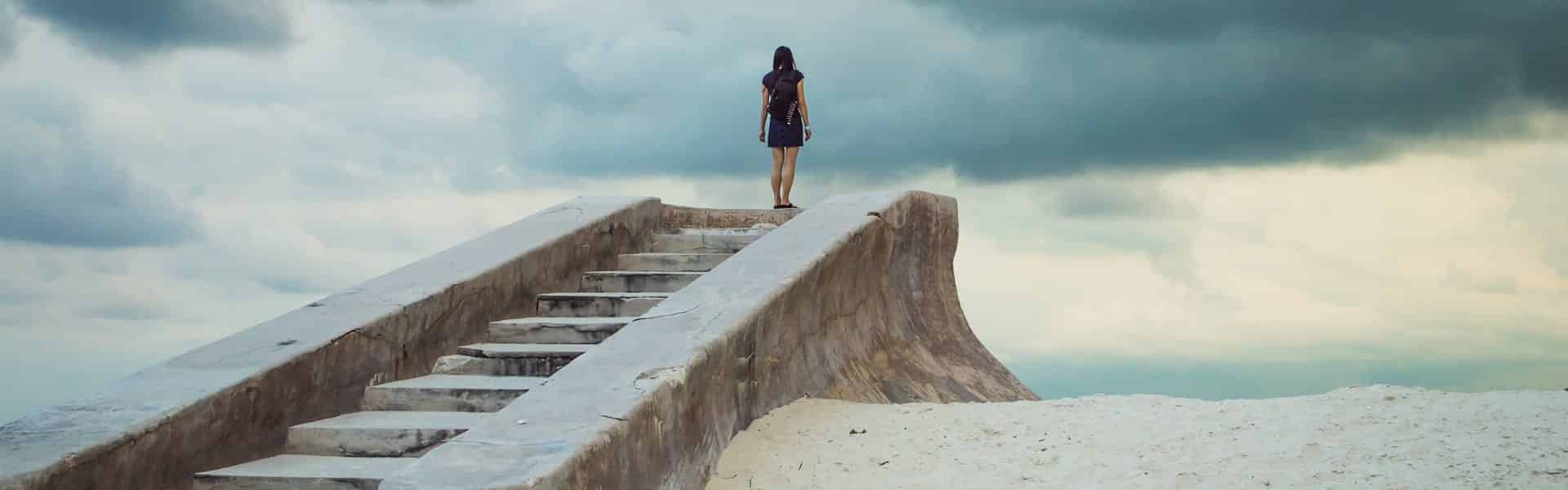 Back of woman who has climbed up stairs and is standing on platform with dark clouds ahead o her