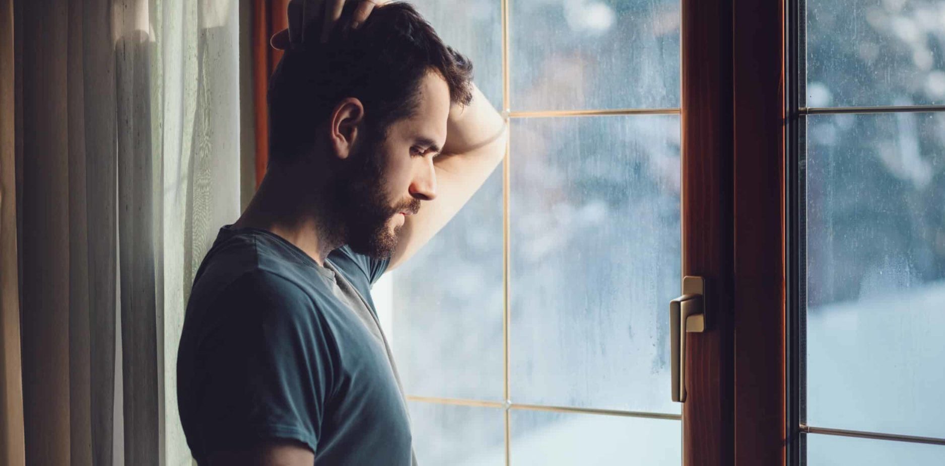 Man leaning against large windows, staring down with hand on his head in need of drug addiction treatment