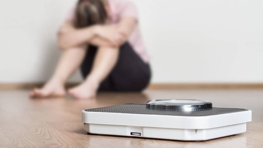 A person sitting head down on the floor is in the background of a weight scale representing the relationship between eating disorders and addiction. 