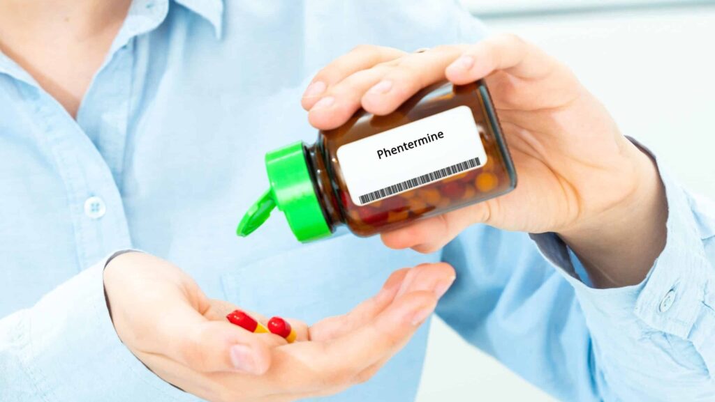A person in a light blue shirt is holding a bottle of Phentermine and pouring out the pills into the other hand. This represents phentermine addiction.