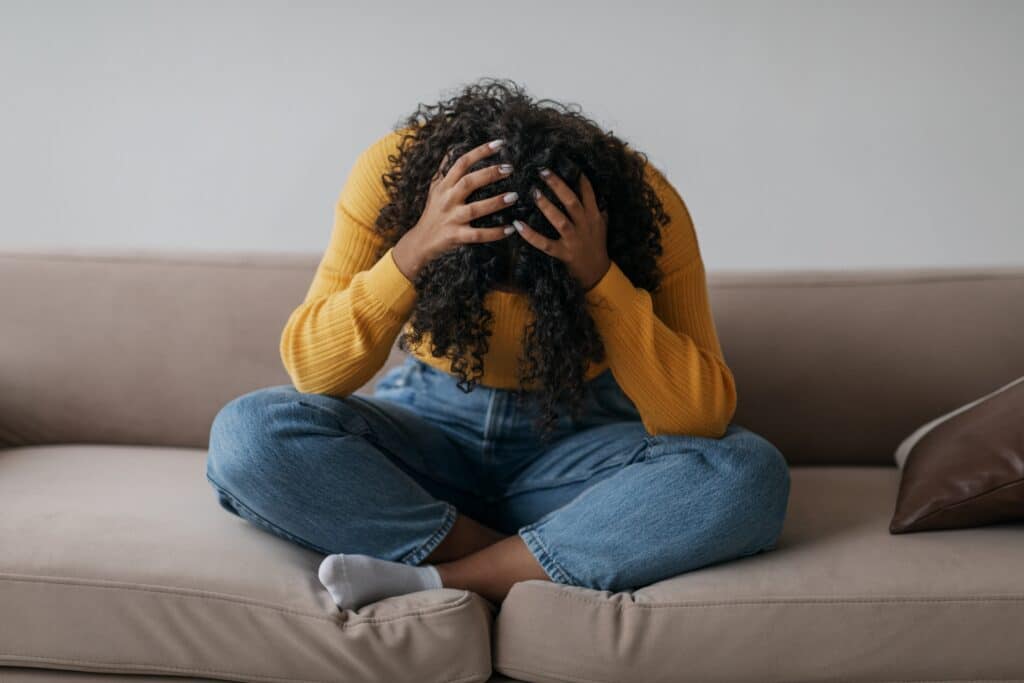 A young woman sitting on a sofa with her hands on her bent down head is an ideal candidate for ketamine treatment for PTSD.