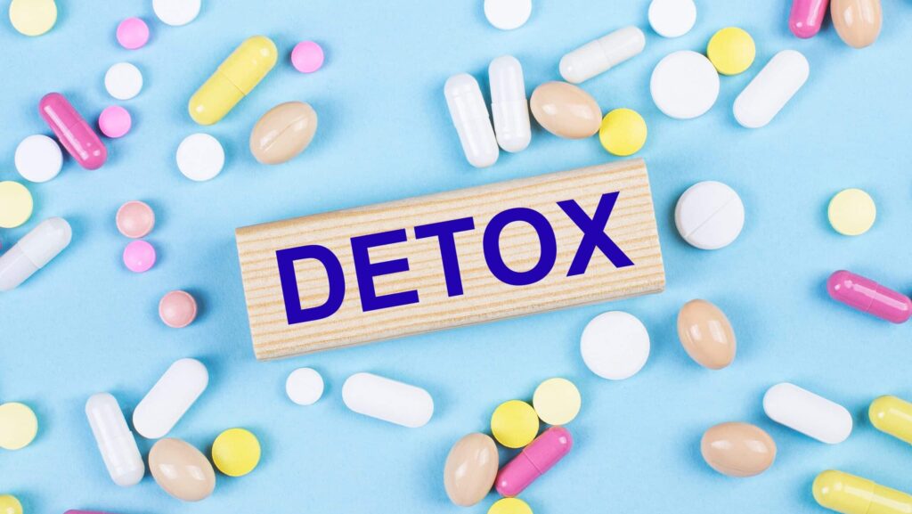 On a light blue background, the word detox printed on a wood block is surrounded by multicolored pills represents how to detox from meth.