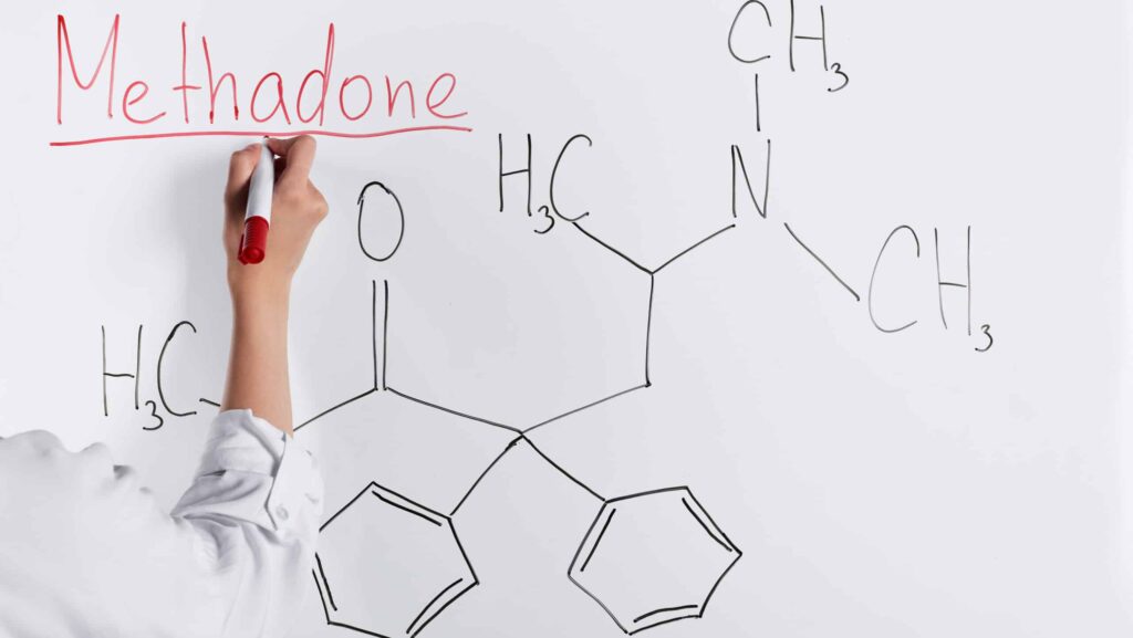 A woman in a white shirt writes the word methadone and draws its chemical structure on a white board to discuss methadone overdose symptoms.