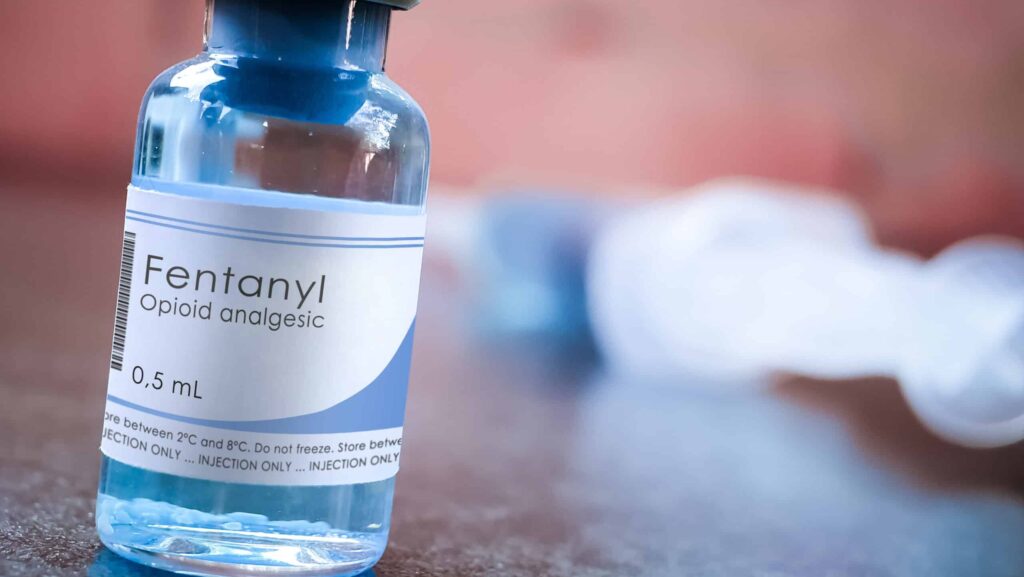 On a blurred red background, a blue-ish glass bottle of liquid fentanyl sits on a table. Users question how long does fentanyl stay in the system?