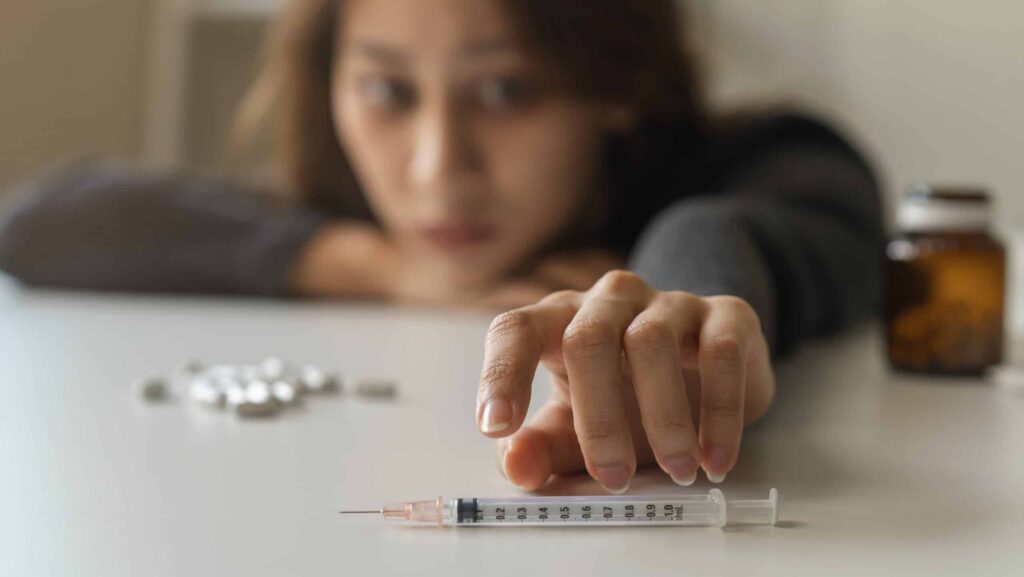 A female young adult taking Xanax and cocaine lays her head on the table with pills while she reaches for a syringe.