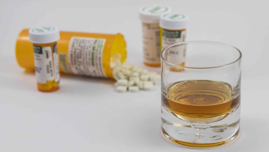 A few prescription bottles and spilled over pills of hydrocodone and alcohol are laid out on a white table.