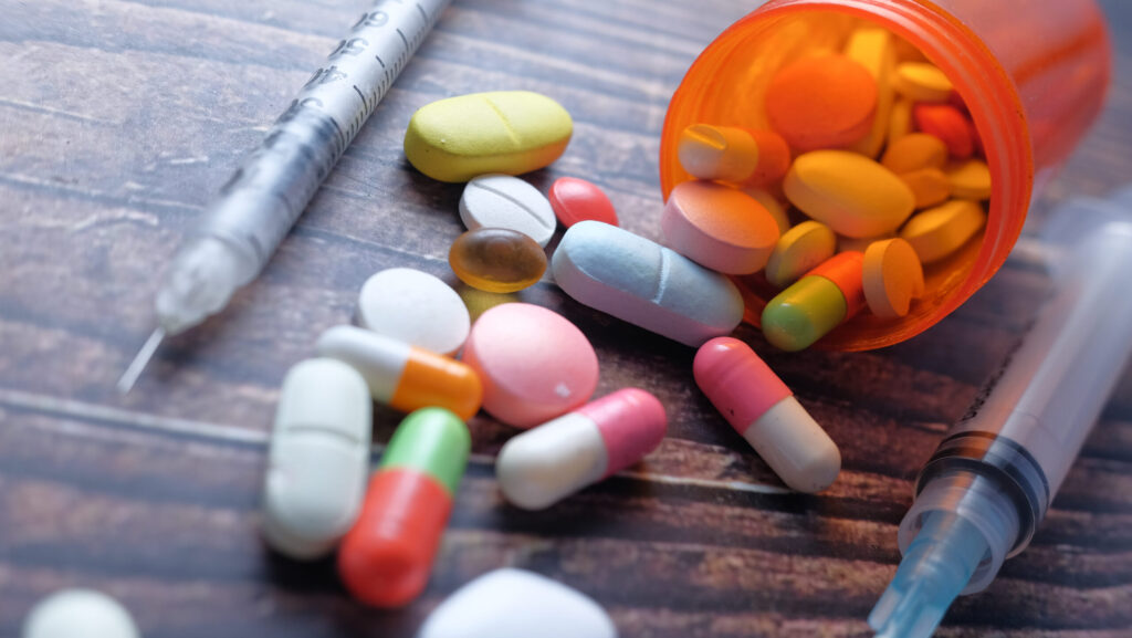 On a wooden table, a spilled over bottle with color pills and tablets lay net to two syringes represents the question of how long does it take to recover from an overdose.