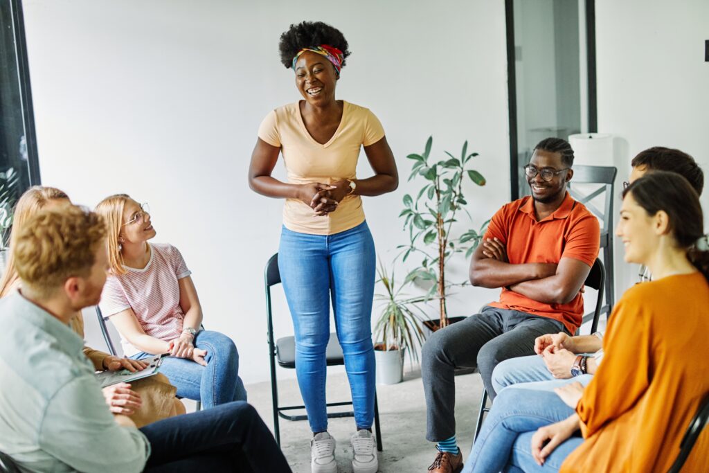 A group of diverse young adults sit in a circle during a PHP or IOP group session while one young women stands to speak about her addiction.