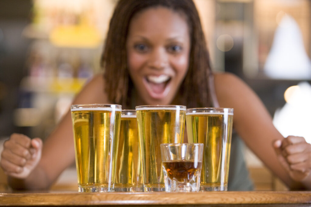 A young woman who is binge drinking is looking at 4 large glasses of beer and one small shot glass of liquor.