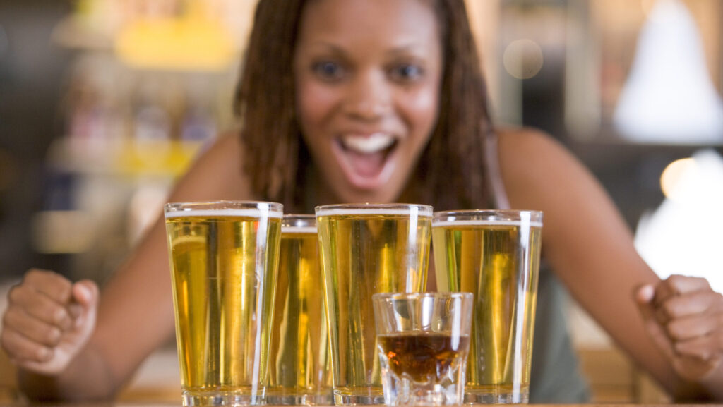 A young woman who is binge drinking is looking at 4 large glasses of beer and one small shot glass of liquor.