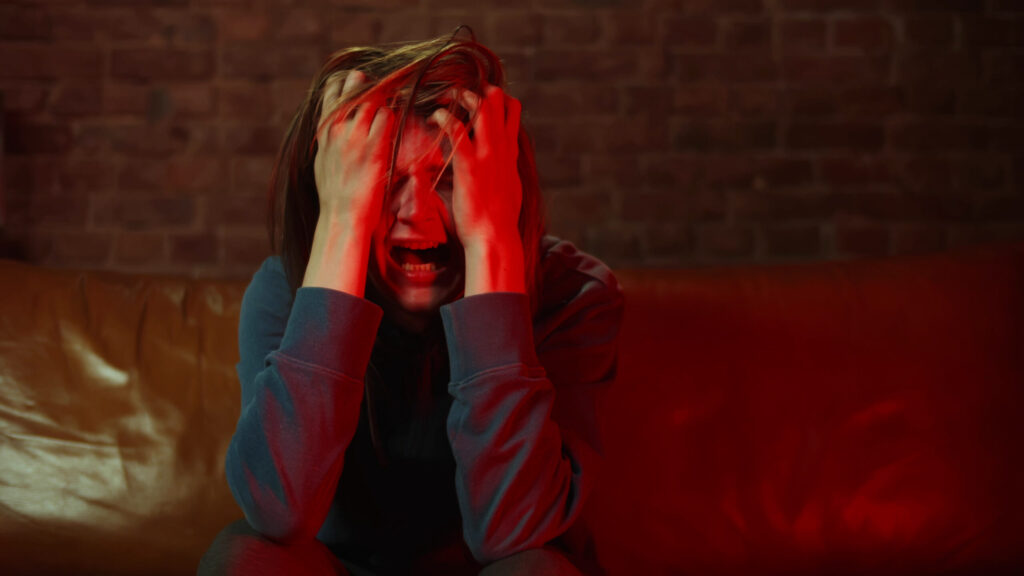 A young man sitting on a leather sofa has his hands to his face in agony because he is experiencing precipitated withdrawal symptoms.