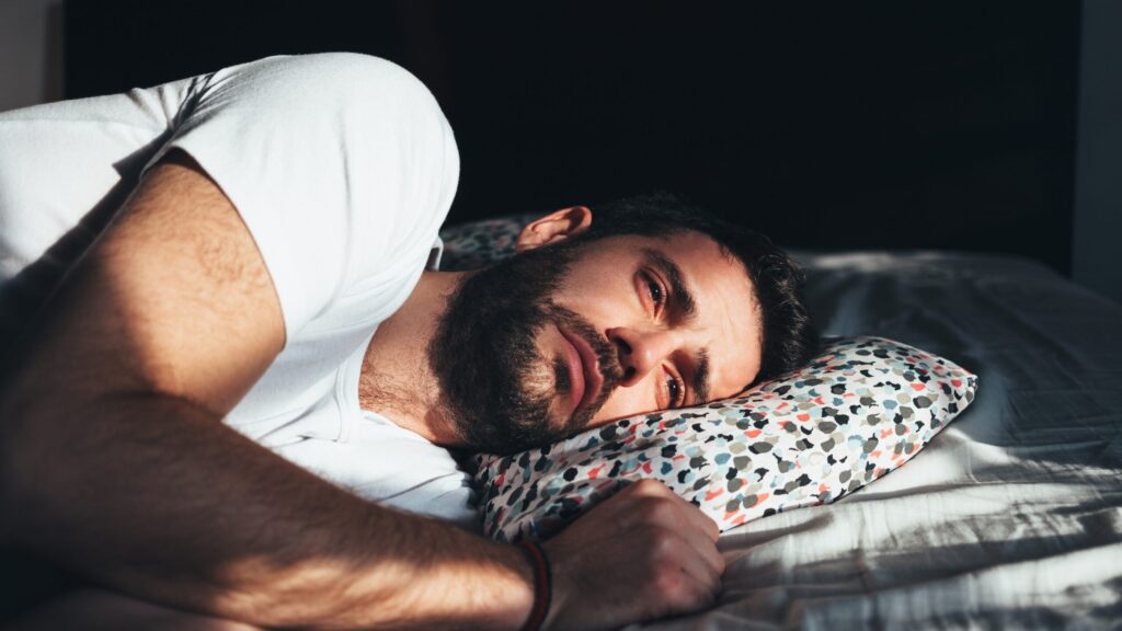 A man with bipolar depression and alcohol addiction is laying on the side of his bed experiencing negative side effects of both diseases.