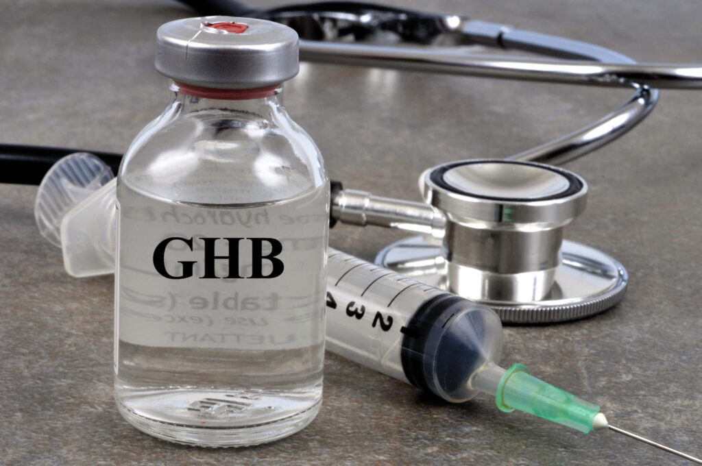 A glass vial of GHB sits next to a syringe and stethoscope represents the side effects you can experience with withdrawal from GHB.