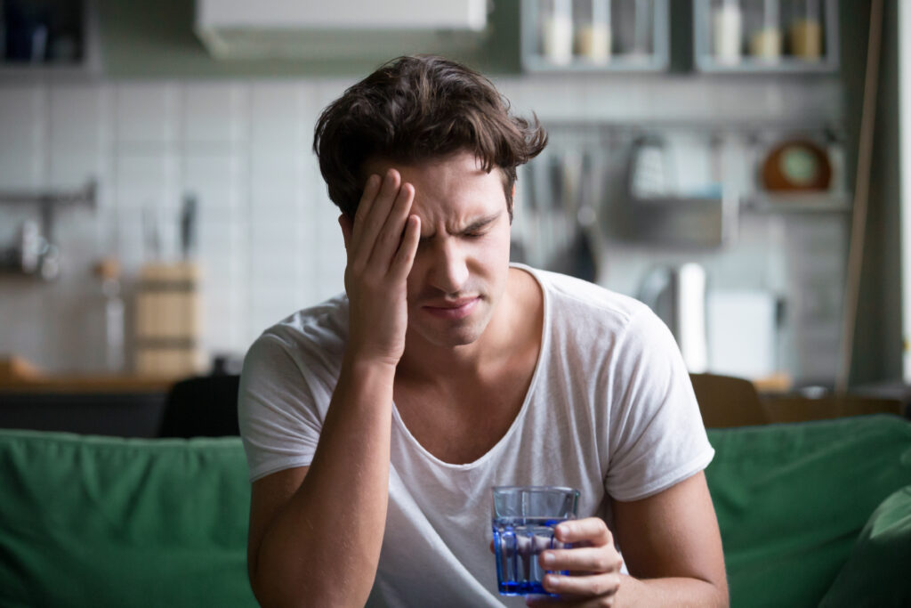 A young man in a white shirt is holding a cup of water with his hands on his temples because he is experiencing a headache from Seroquel withdrawal.