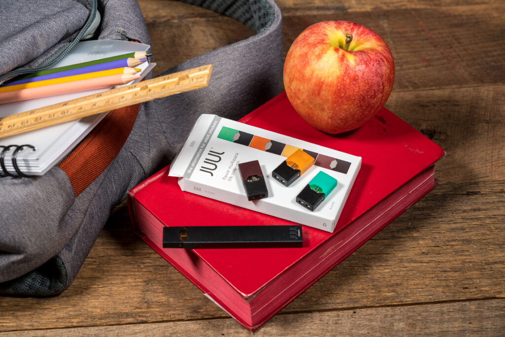 An open backpack with books, color pencils and a ruler is laid next to a book with an apple and a Juul e-cigarette stacked on top.