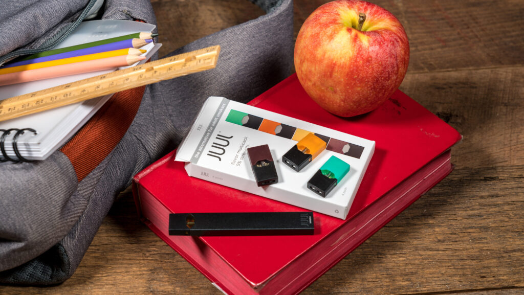 An open backpack with books, color pencils and a ruler is laid next to a book with an apple and a Juul e-cigarette stacked on top.