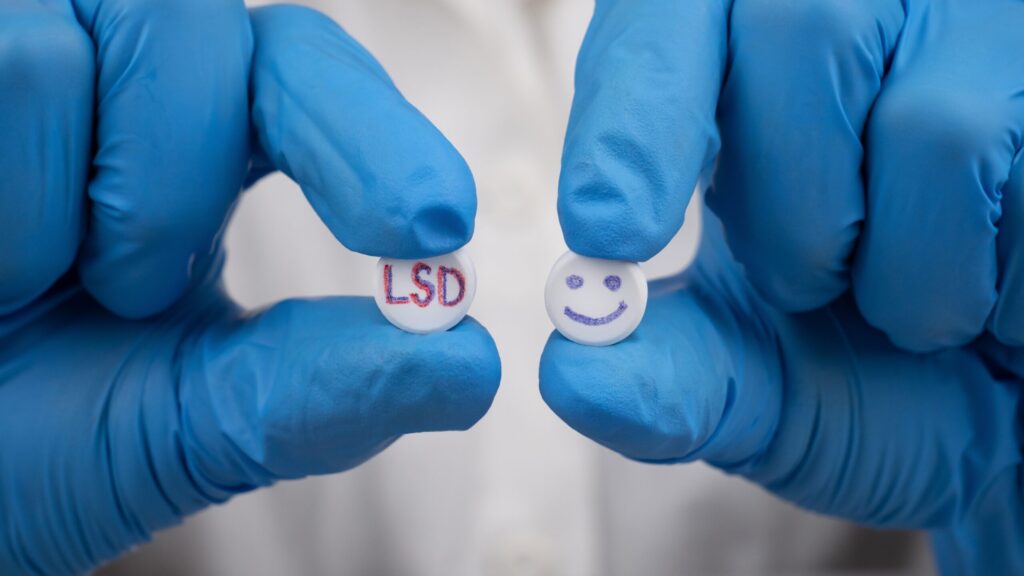 A person wearing blue gloves holding up two different LSD pills, one with the letters LSD and the other with a smiley face.