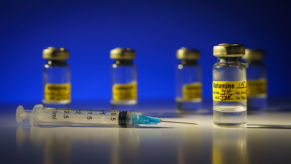 Multiple glass vials of ketamine are next to a syringe represents how long ketamine pain relief can last after initial therapy session.