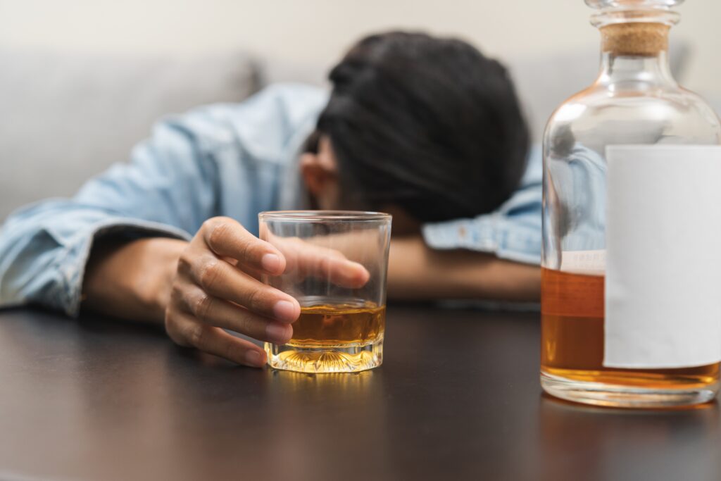 A young man with his head down on his arm holds a glass of alcohol on a table represents ethanol abuse.