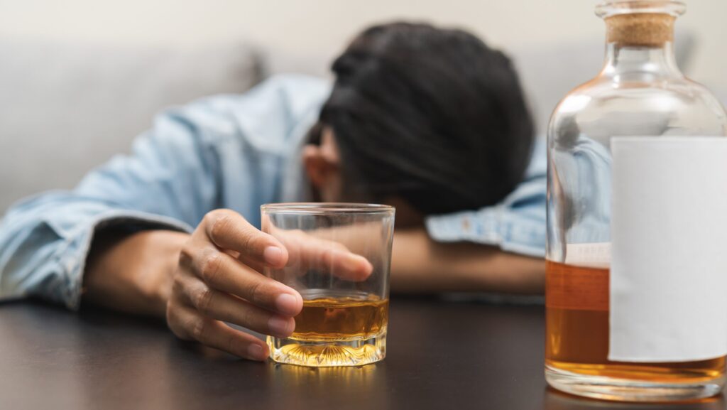 A young man with his head down on his arm holds a glass of alcohol on a table represents ethanol abuse.