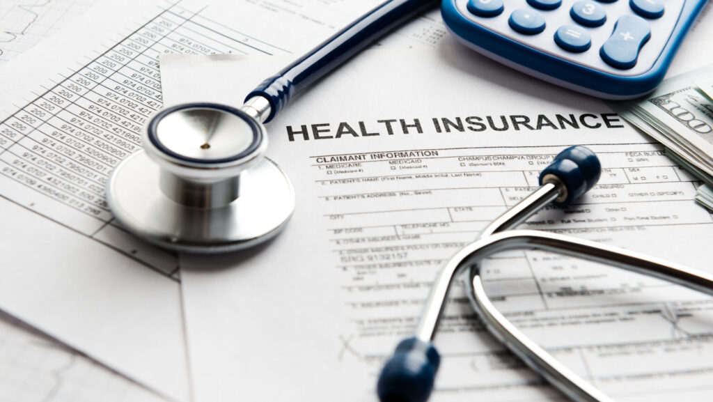 Medical insurance papers lay on a desk with a stethoscope, money and calculator.