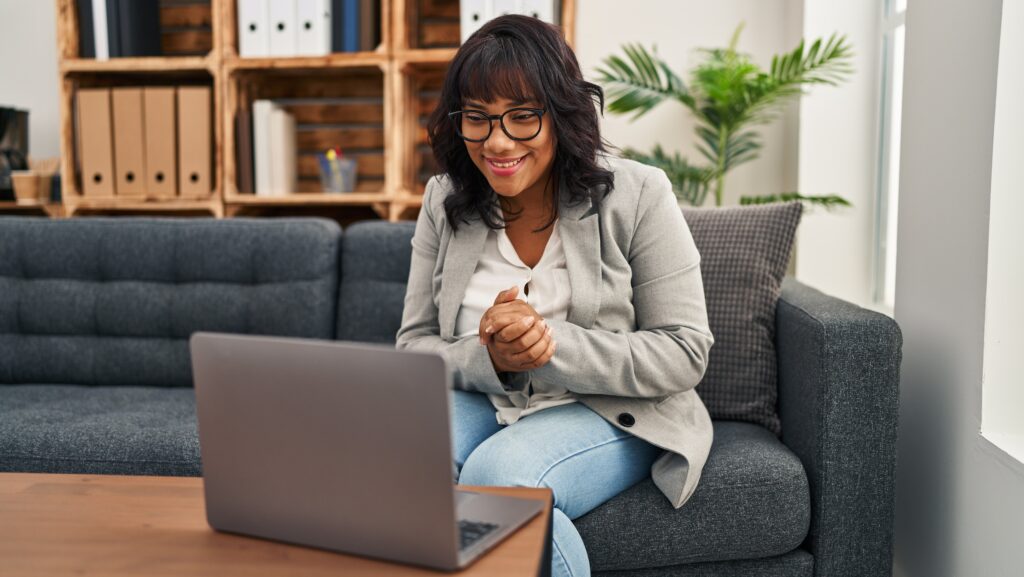 A young women with glasses sitting on the sofa on her laptop engaging in online rehab therapy in the comforts of her home.