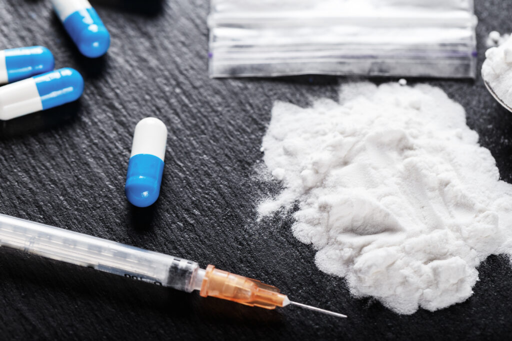 A white powder representing crushed up tramadol, blue and white tablets and a syringe lay on a black table represent a person snorting tramadol.