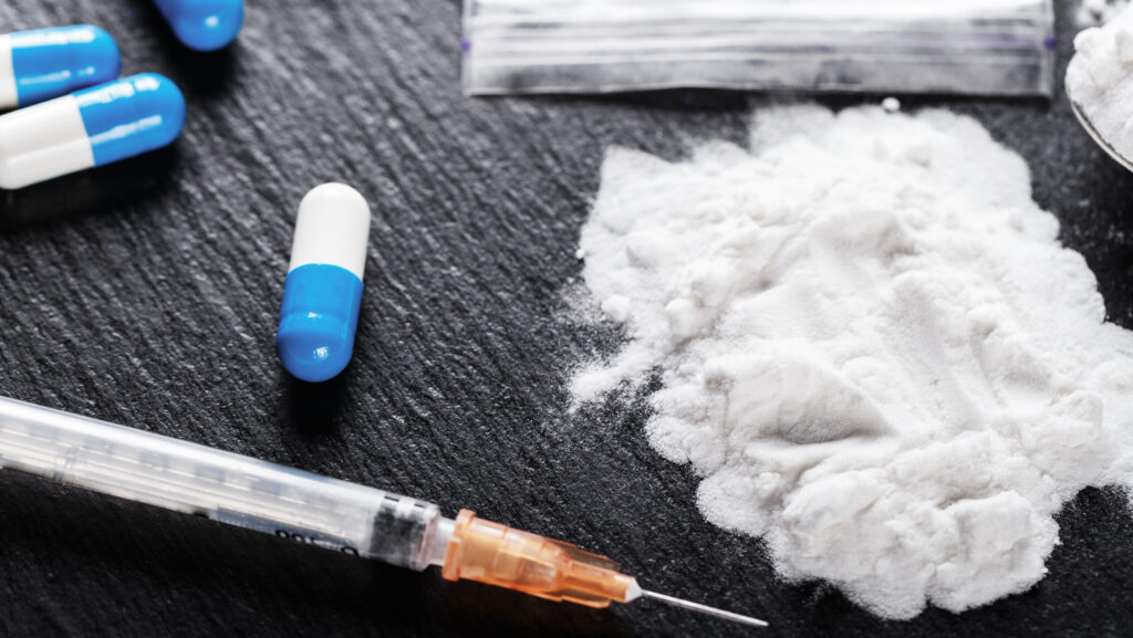 A white powder representing crushed up tramadol, blue and white tablets and a syringe lay on a black table represent a person snorting tramadol.