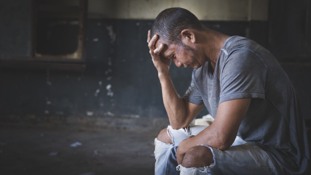 A man in grey shirt and ripped jeans going through withdrawal symptoms you can die from is sitting uncomfortably with his hand covering his face.