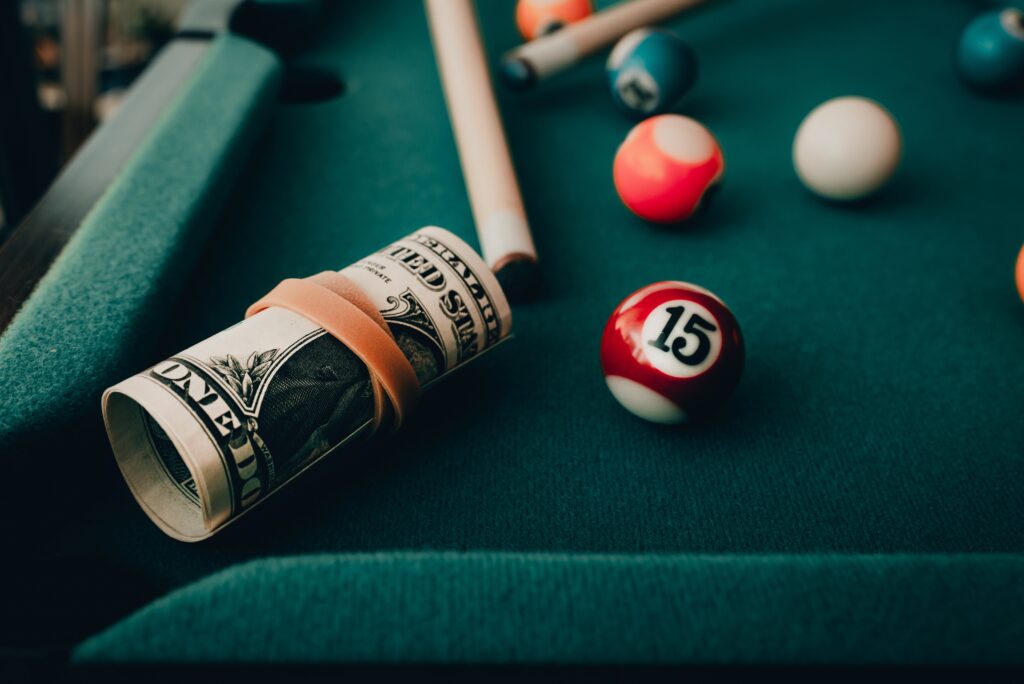 On a billards table with a few balls, there is rolled up wad of cash representing a gambling and substance use disorder cross addiction.