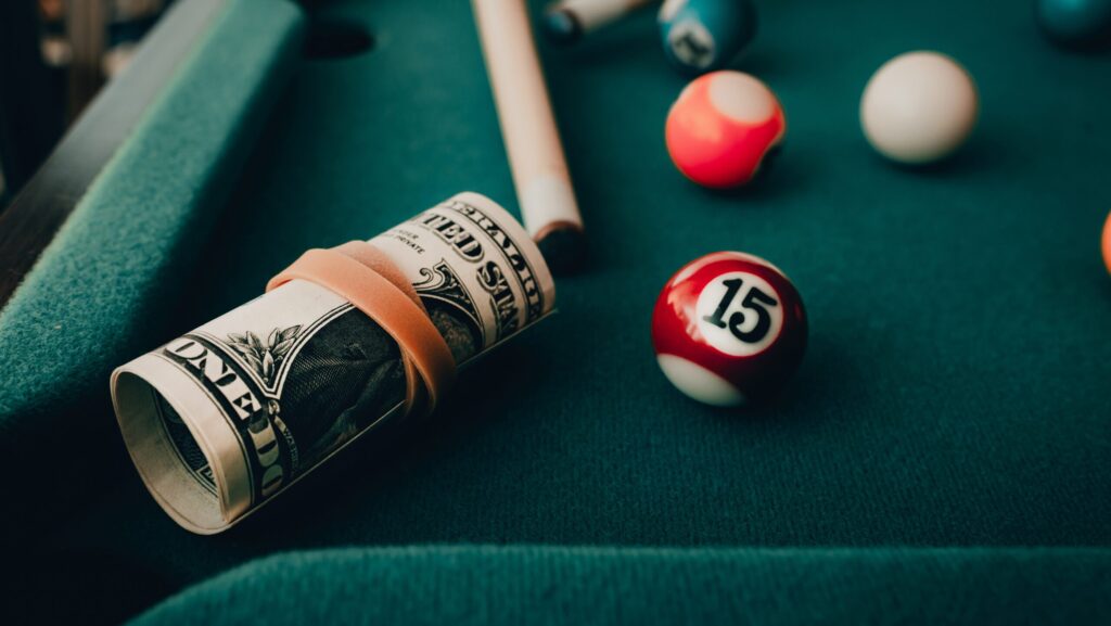 On a billards table with a few balls, there is rolled up wad of cash representing a gambling and substance use disorder cross addiction.