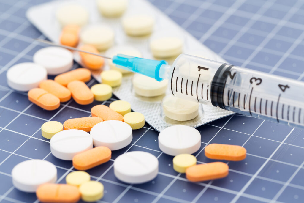 A tray of pills and tablets of various shapes and sizes next to a syringe represents types of medication assisted treatment (MAT).