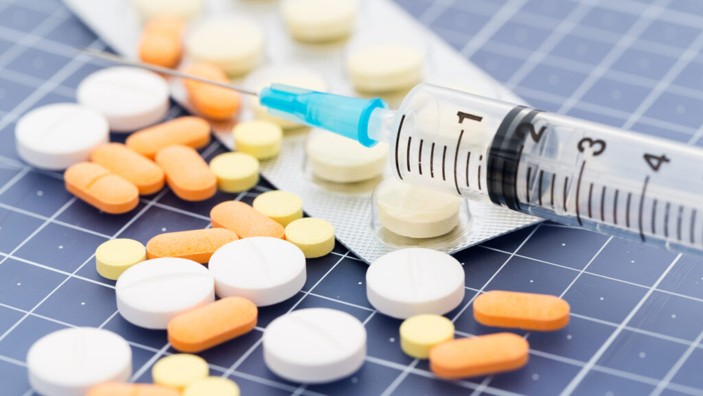 A tray of pills and tablets of various shapes and sizes next to a syringe represents types of medication assisted treatment (MAT).