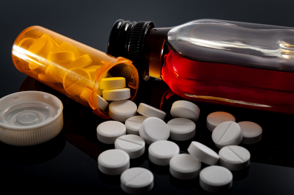 An orange bottle of opioid pills spills over next to a closed bottle of an amber liquid substance represents k18 pills.