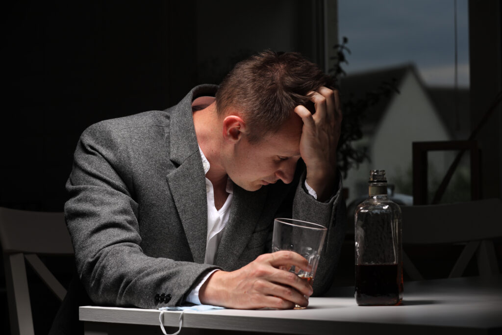 A man in a grey suit sitting alone with his head in his hand and holding a glass of alcohol in the other hand.