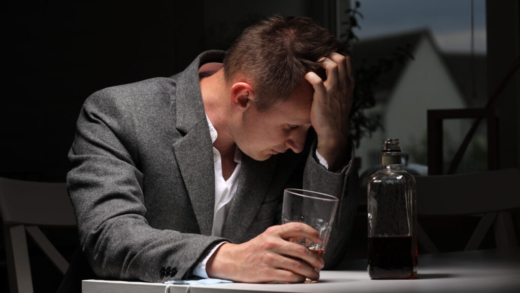 A man in a grey suit sitting alone with his head in his hand and holding a glass of alcohol in the other hand.