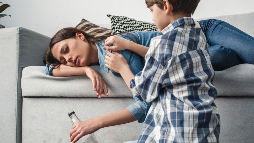 A young boy confronting his mother as she lays on sofa passed out with a bottle of alcohol in her hand.