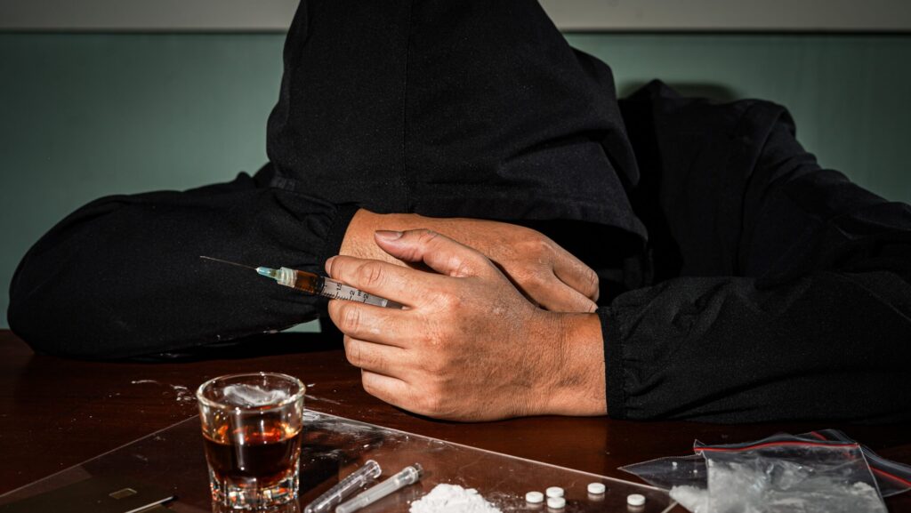 A man in black hoodie going through the 5 stages of addiction is holding a syringe in front of table of drugs and alcohol.