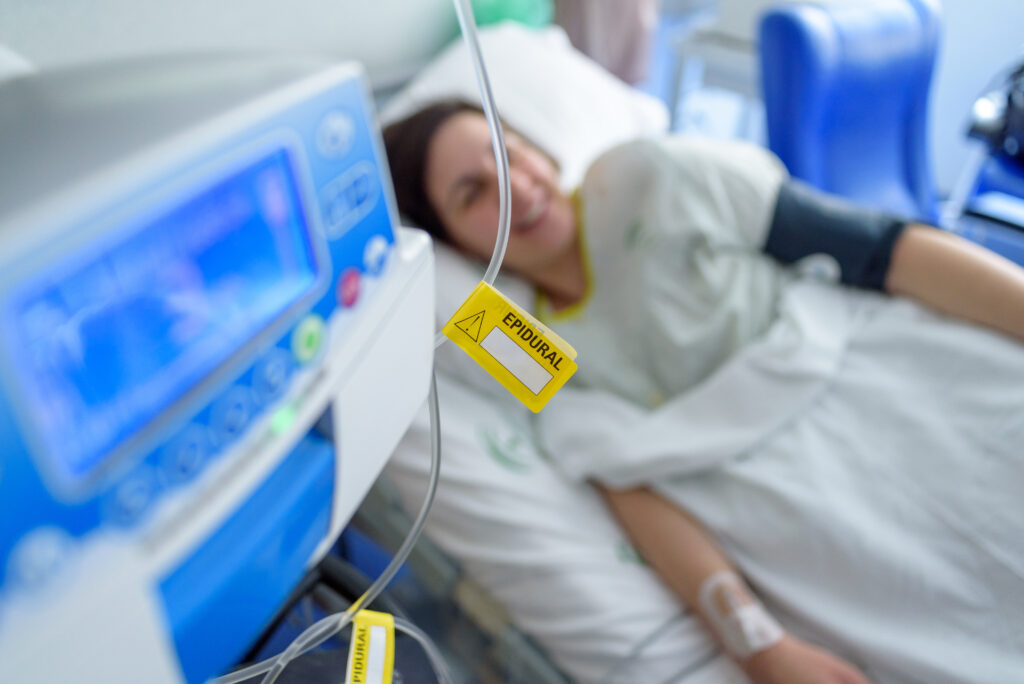 In a hospital setting a patient is laying in bed with an attached epidural IV line hanging near a machine.