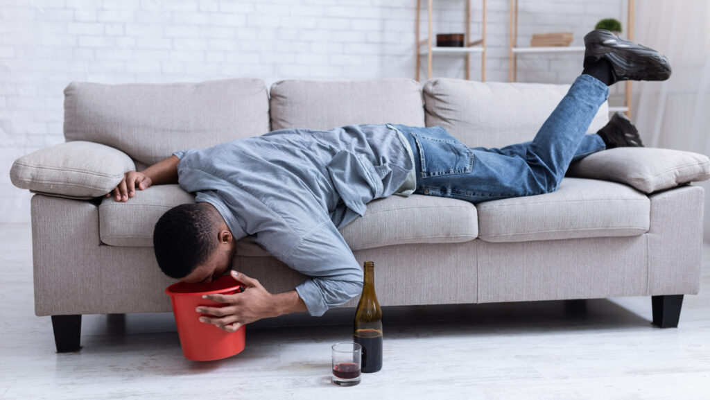 A man laying on the sofa with his head over a bucket next to a bottle of wine is experiencing alcohol poisoning.