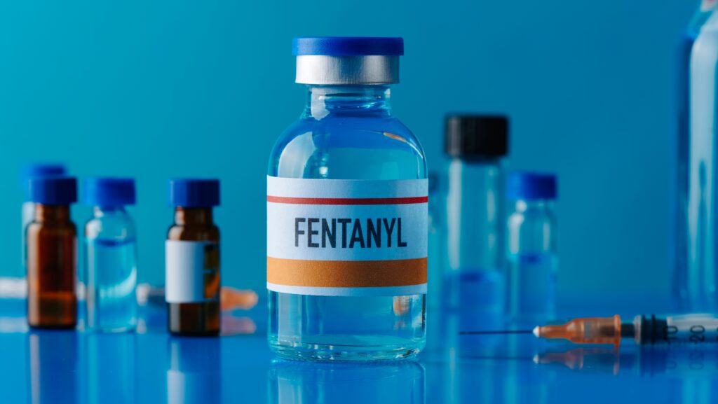 A glass bottle of fentanyl sits on a table with other glass vials and a syringe in the background.