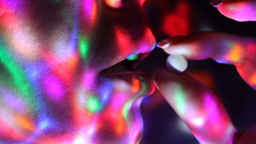 A close up of a young woman holding up a small round ecstasy pill next to her mouth and putting her self at risk for OD on Ecstasy.