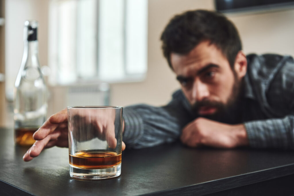 A man with end stage alcoholism reaches for a glass filled with alcohol as he slumps over a table.