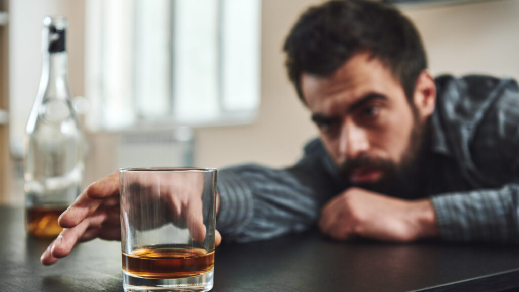 A man with end stage alcoholism reaches for a glass filled with alcohol as he slumps over a table.