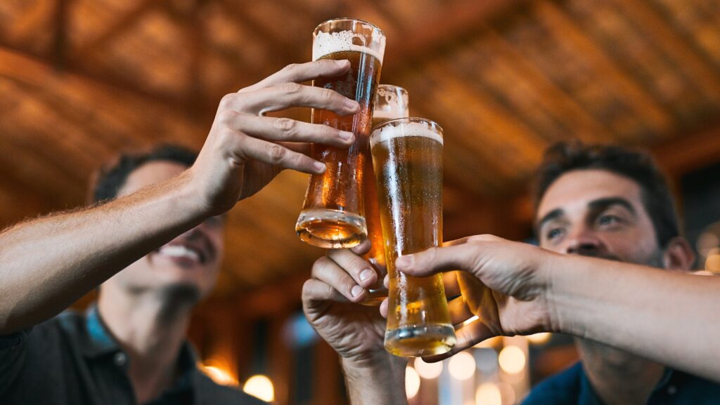 A group of young men holding up tall glasses of beers and unaware of the relationship between alcohol and prostate cancer.